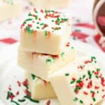 Three square pieces of Sugar Cookie Fudge stacked on a plate, topped with red and green holiday sprinkles, with more fudge pieces in the background.