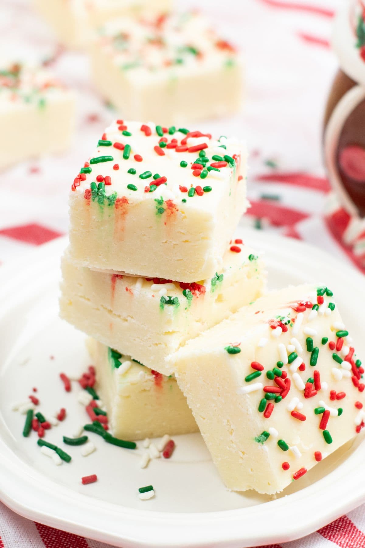 Three square pieces of Sugar Cookie Fudge stacked on a plate, topped with red and green holiday sprinkles, with more fudge pieces in the background.
