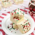 Three pieces topped with red and green sprinkles are stacked on a white plate, with more fudge and a mug in the background on a striped cloth.