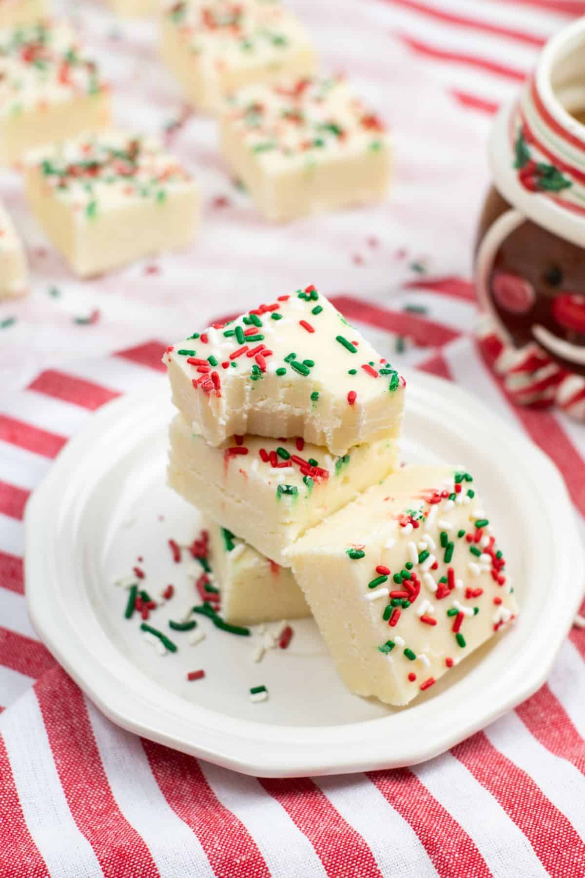 Three pieces topped with red and green sprinkles are stacked on a white plate, with more fudge and a mug in the background on a striped cloth.