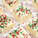 Squares of Sugar Cookie Fudge topped with red, green, and white sprinkles are arranged on striped parchment paper.