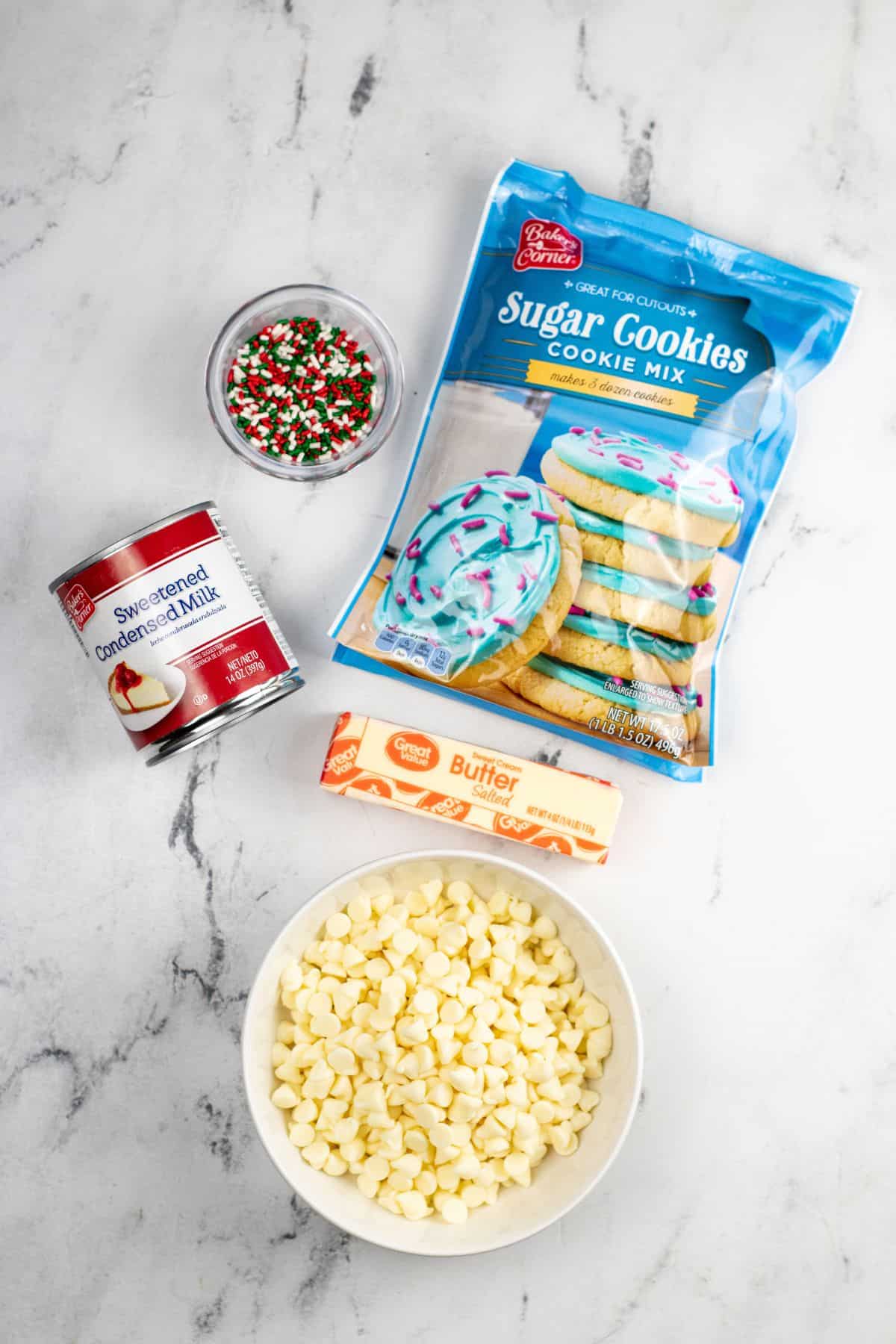 Ingredients include sugar cookie mix, a stick of butter, a bowl of white chocolate chips, sweetened condensed milk, and holiday sprinkles—all set on a marble surface.