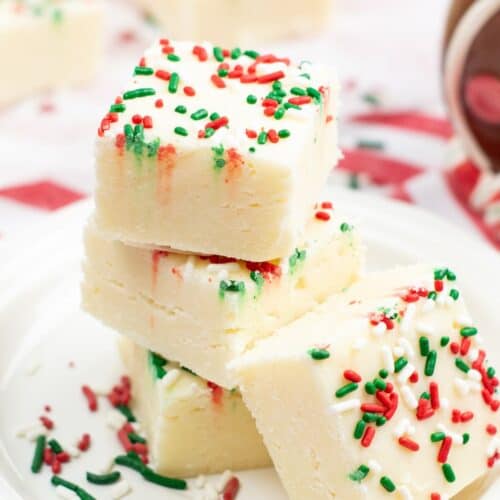 Three pieces of Sugar Cookie Fudge stacked on a plate, topped with red, green, and white sprinkles, with more fudge and festive sprinkles in the background.