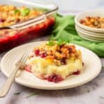 A serving of Thanksgiving Leftovers Casserole with mashed potatoes, cranberries, and stuffing on a white plate with a fork; a casserole dish and stacked bowls are in the background.