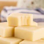 A stack of white chocolate fudge squares, with the top piece showing bite marks—this irresistible treat is perfect for anyone seeking a classic vanilla fudge recipe. A striped cloth is blurred in the background.