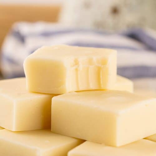 A stack of white chocolate fudge squares, with the top piece showing bite marks—this irresistible treat is perfect for anyone seeking a classic vanilla fudge recipe. A striped cloth is blurred in the background.