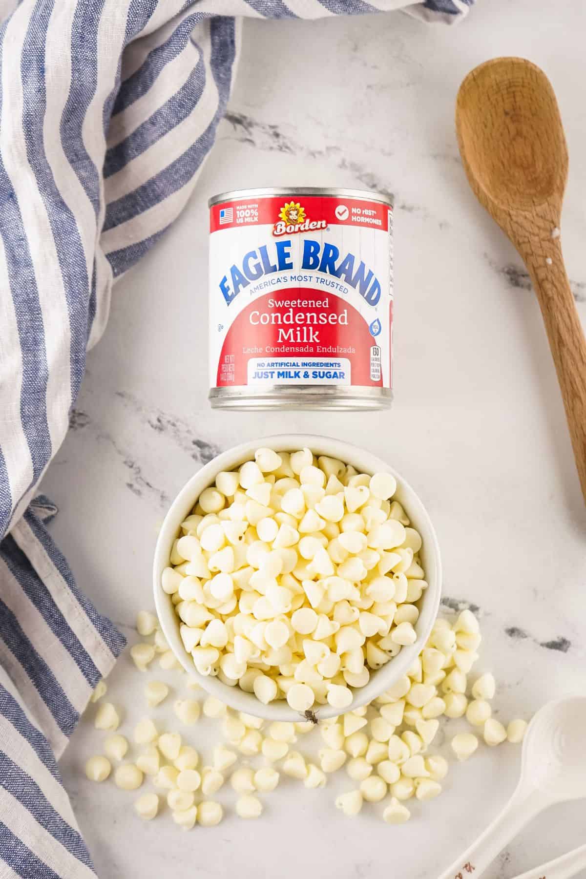 A can of Eagle Brand sweetened condensed milk and a bowl of white chocolate chips, with a wooden spoon and striped cloth nearby.