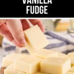 A hand holds a square piece of vanilla fudge above a pile of similar fudge squares on a wooden board, with white chocolate chips nearby—perfect inspiration for your next vanilla fudge recipe.