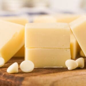 Two stacked pieces of white chocolate fudge, made from a creamy vanilla fudge recipe, are surrounded by white chocolate chips on a wooden surface.