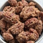 A bowl filled with Cinnamon Roasted Pecans, each coated in a sweet layer of sugar and spices, sits on a light-colored surface.