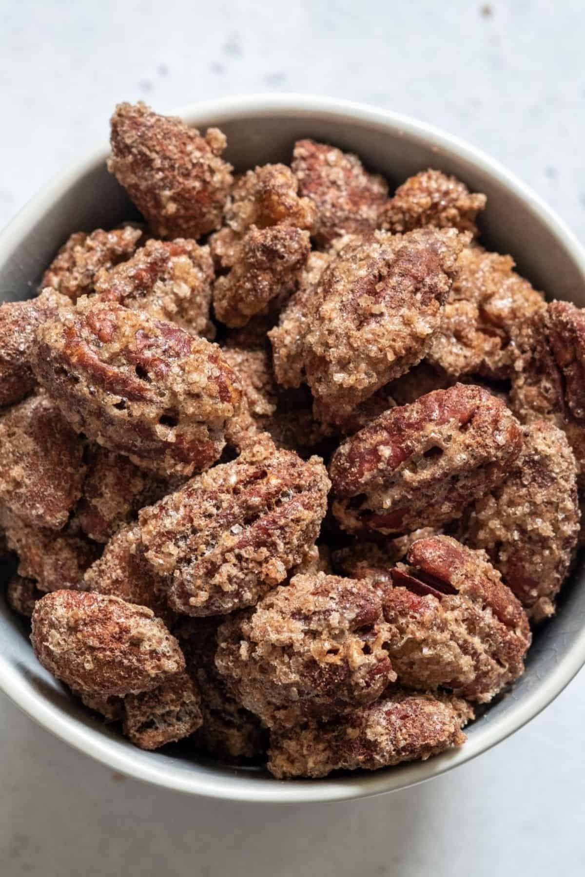 A bowl filled with Cinnamon Roasted Pecans, each coated in a sweet layer of sugar and spices, sits on a light-colored surface.
