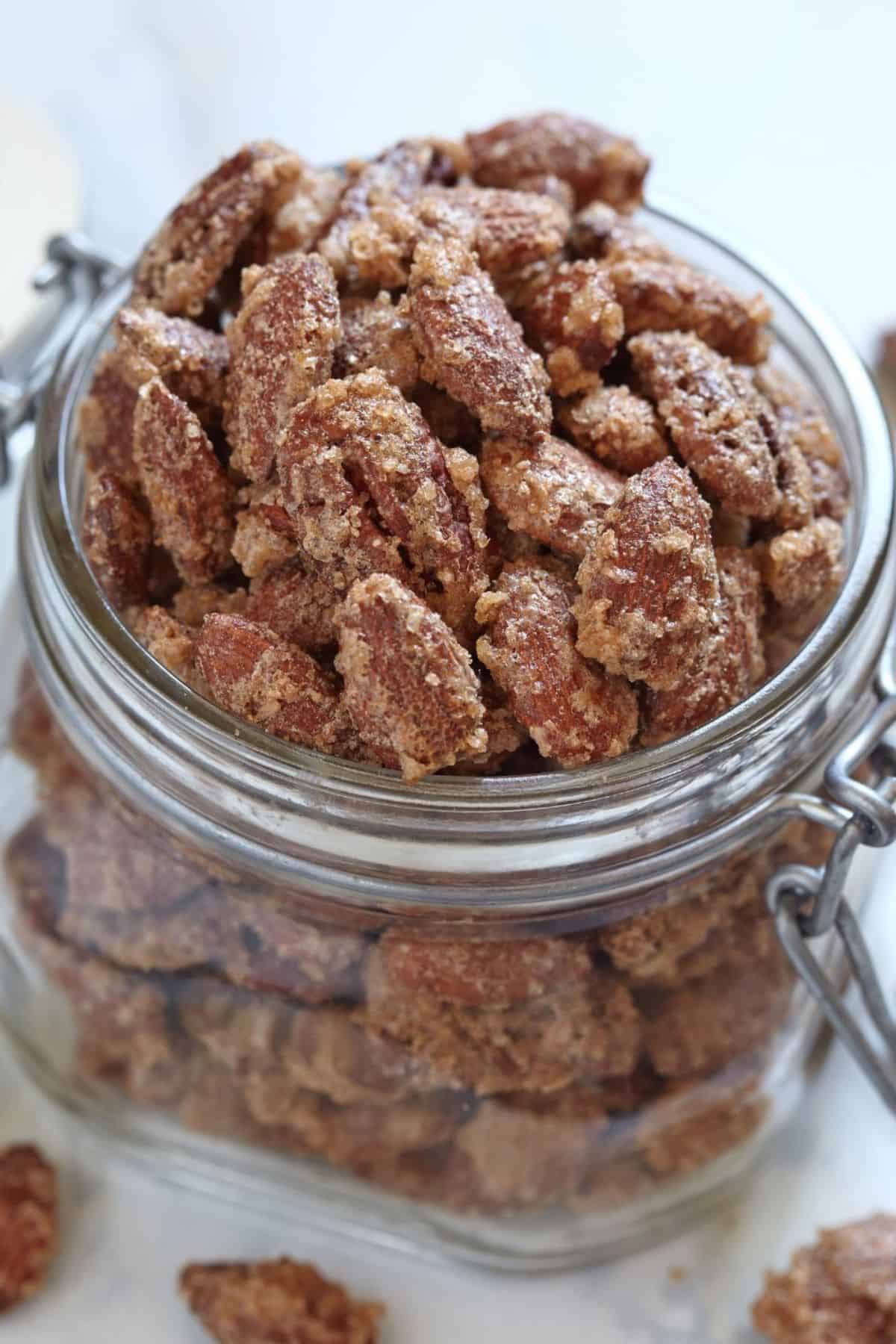 A glass jar filled with nuts, their sugar-coated surfaces glistening, shown in close-up on a light background.