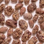 A variety of candied nuts, including cinnamon roasted pecans and almonds, coated in a sugar and spice mixture, spread out on a light pink surface.