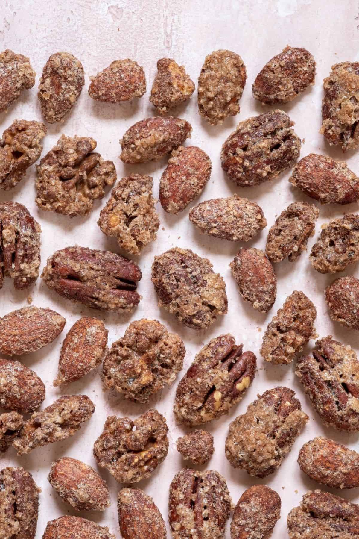 A variety of candied nuts, including cinnamon roasted pecans and almonds, coated in a sugar and spice mixture, spread out on a light pink surface.
