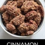 A white bowl filled with Cinnamon Roasted Pecans, each nut coated in a brown, textured cinnamon-sugar mixture, sits on a light surface.
