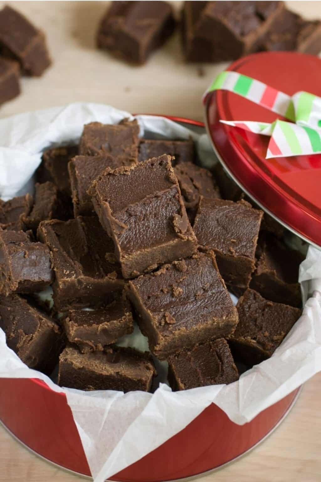 A red tin lined with parchment paper is filled with squares of Easy Microwave Fudge, the lid partially open and decorated with a striped ribbon.