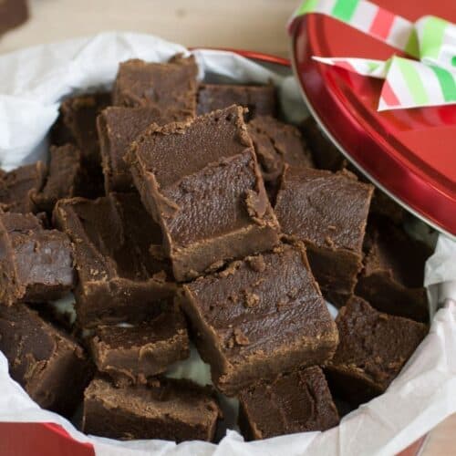 A red tin lined with parchment paper is filled with squares of Easy Microwave Fudge, the lid partially open and decorated with a striped ribbon.