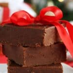 Three pieces of Easy Microwave Fudge stacked on a plate, tied together with a red ribbon bow, with festive decorations blurred in the background.