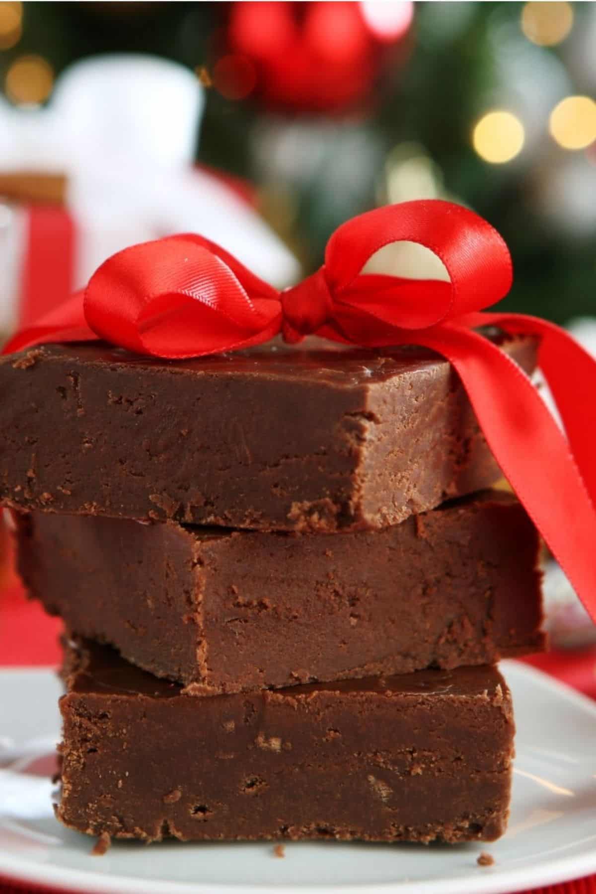 Three pieces of Easy Microwave Fudge stacked on a plate, tied together with a red ribbon bow, with festive decorations blurred in the background.