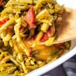 A wooden spoon holds a serving of Green Beans with Bacon and Onion, featuring tender green beans mixed with savory bacon pieces in a white dish.