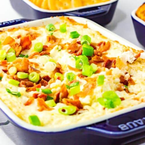 A baking dish of creamy bacon and cheese dip is topped with chopped green onions and bacon bits, with a dish of round crackers in the background.