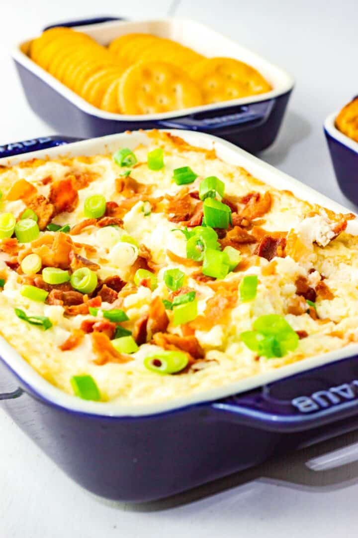 A baking dish of creamy bacon and cheese dip is topped with chopped green onions and bacon bits, with a dish of round crackers in the background.