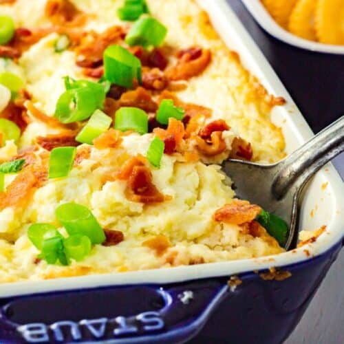 Rectangular baking dish with a creamy baked cheese dip topped with chopped bacon and green onions, with a metal spoon and crackers visible in the background.