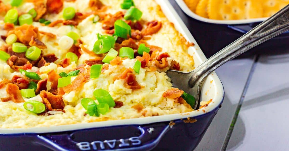 Rectangular baking dish with a creamy baked cheese dip topped with chopped bacon and green onions, with a metal spoon and crackers visible in the background.