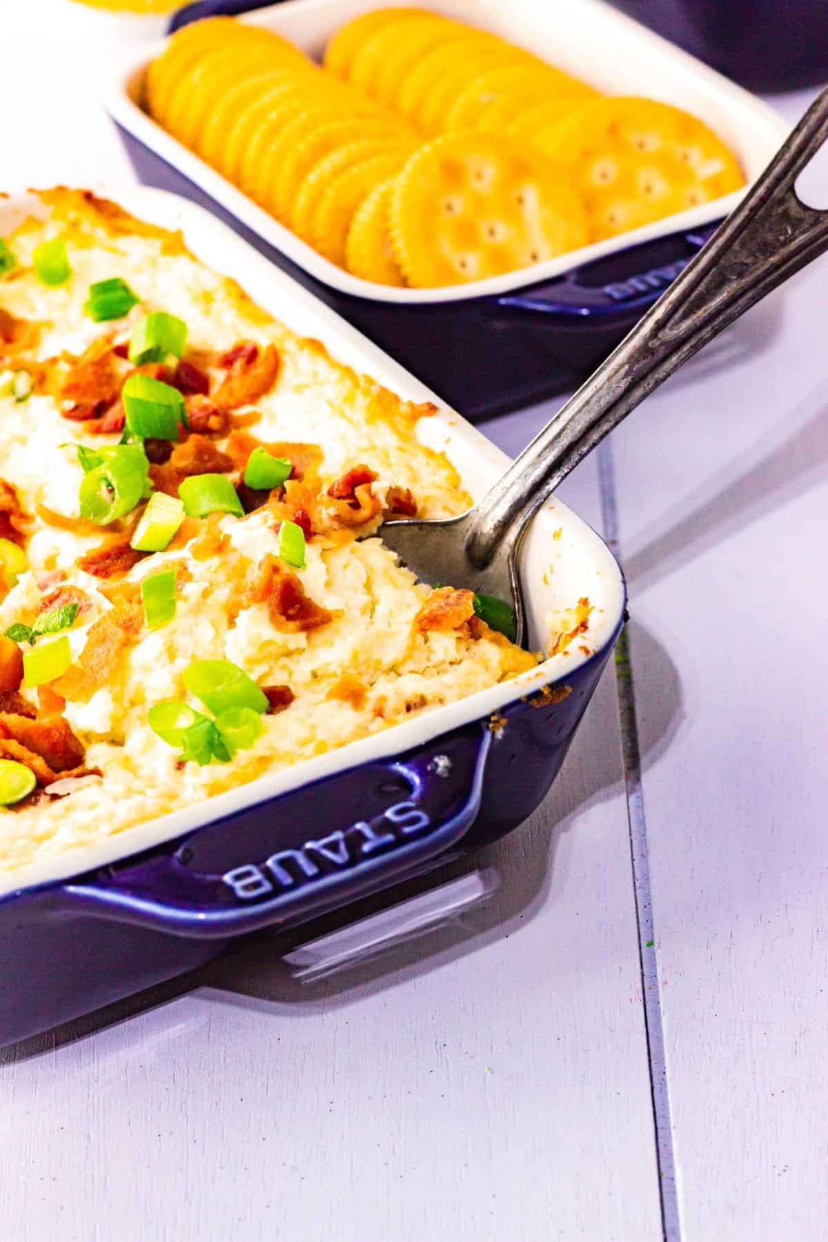 A baked cheese dip topped with green onions and bacon is served in a blue dish next to a tray of round crackers.