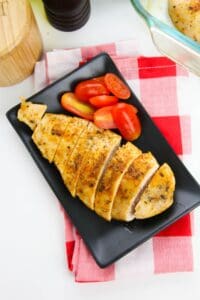 Sliced baked chicken cutlets and halved grape tomatoes arranged on a black rectangular plate, set on a red and white checkered napkin.