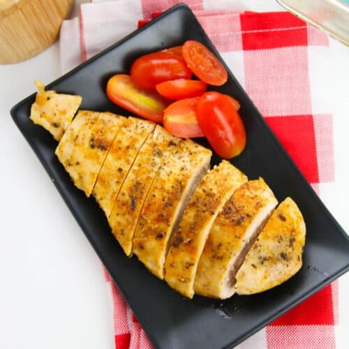 Sliced baked chicken cutlets and halved grape tomatoes arranged on a black rectangular plate, set on a red and white checkered napkin.