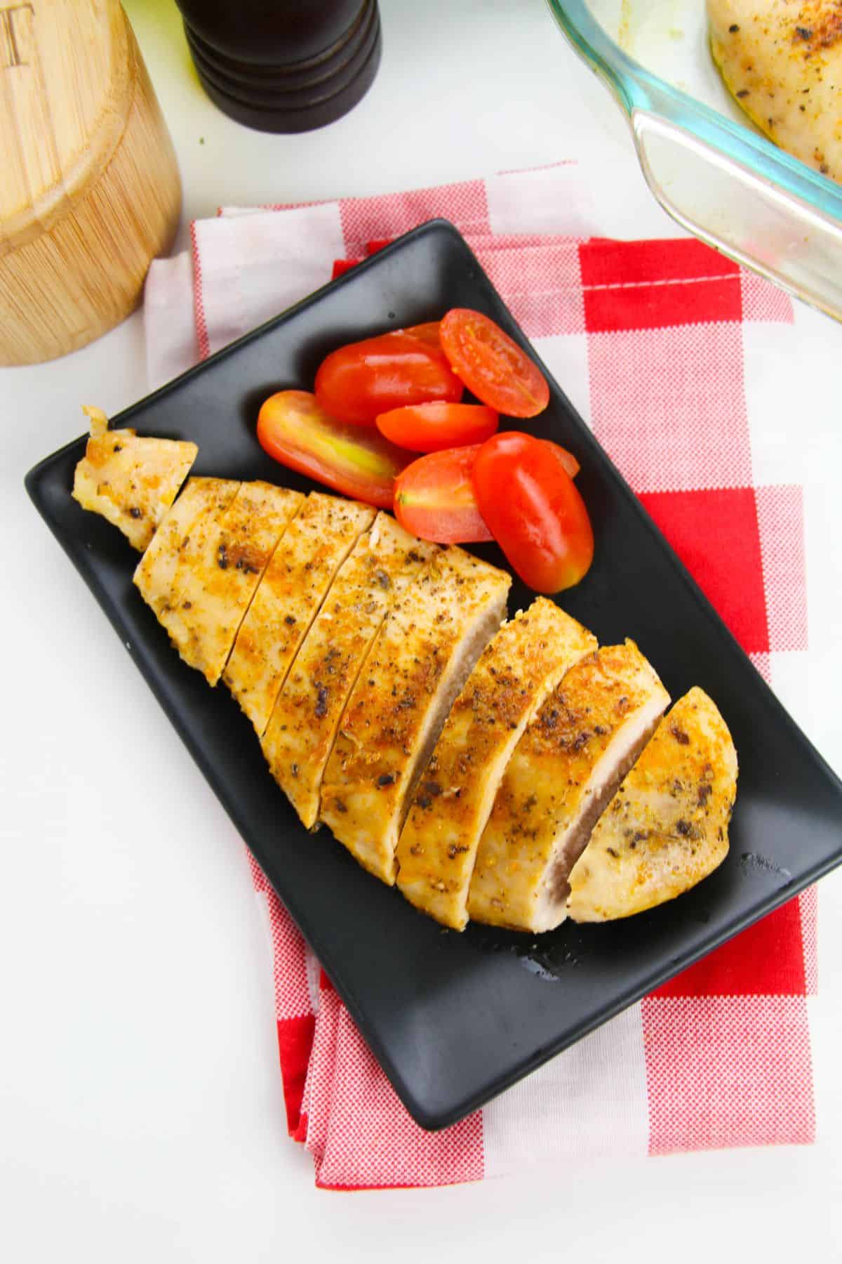 Sliced baked chicken cutlets and halved grape tomatoes arranged on a black rectangular plate, set on a red and white checkered napkin.