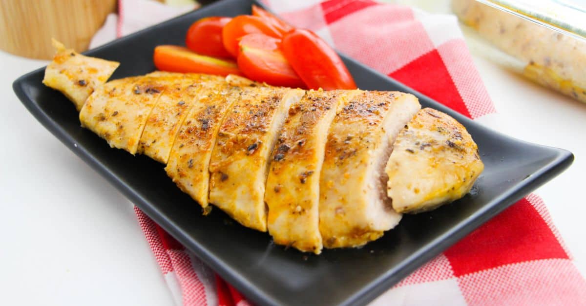 Baked chicken cutlets sliced and served on a black rectangular plate with tomato wedges, placed on a red and white checkered cloth.
