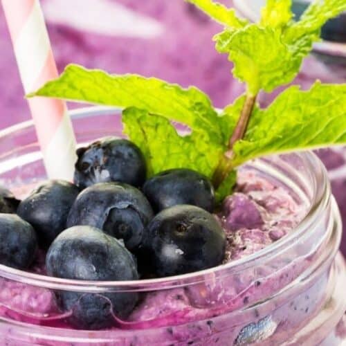 A mason jar filled with blueberry yogurt smoothie, topped with fresh blueberries and a sprig of mint, with a striped straw.
