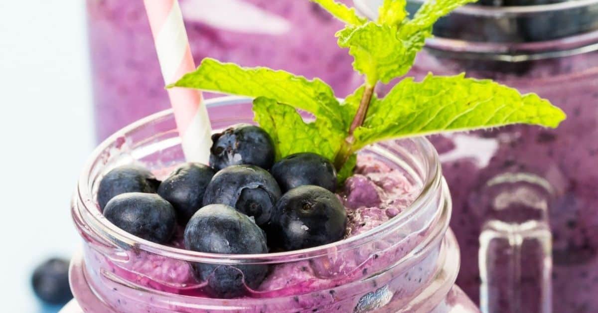 A mason jar filled with blueberry yogurt smoothie, topped with fresh blueberries and a sprig of mint, with a striped straw.
