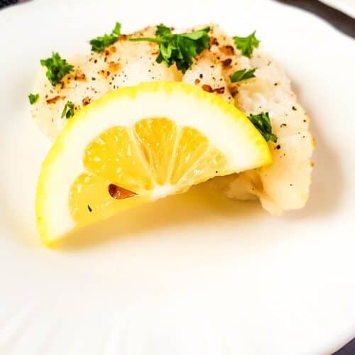 Two pieces of Air Fryer Cod are garnished with chopped parsley, black pepper, and a lemon wedge on a white plate.