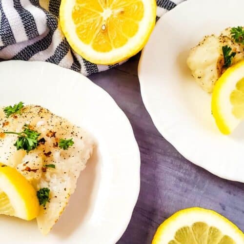 Two white plates with Air Fryer Cod fillets, garnished with lemon slices and parsley, on a gray surface with a striped cloth and additional lemon slices.