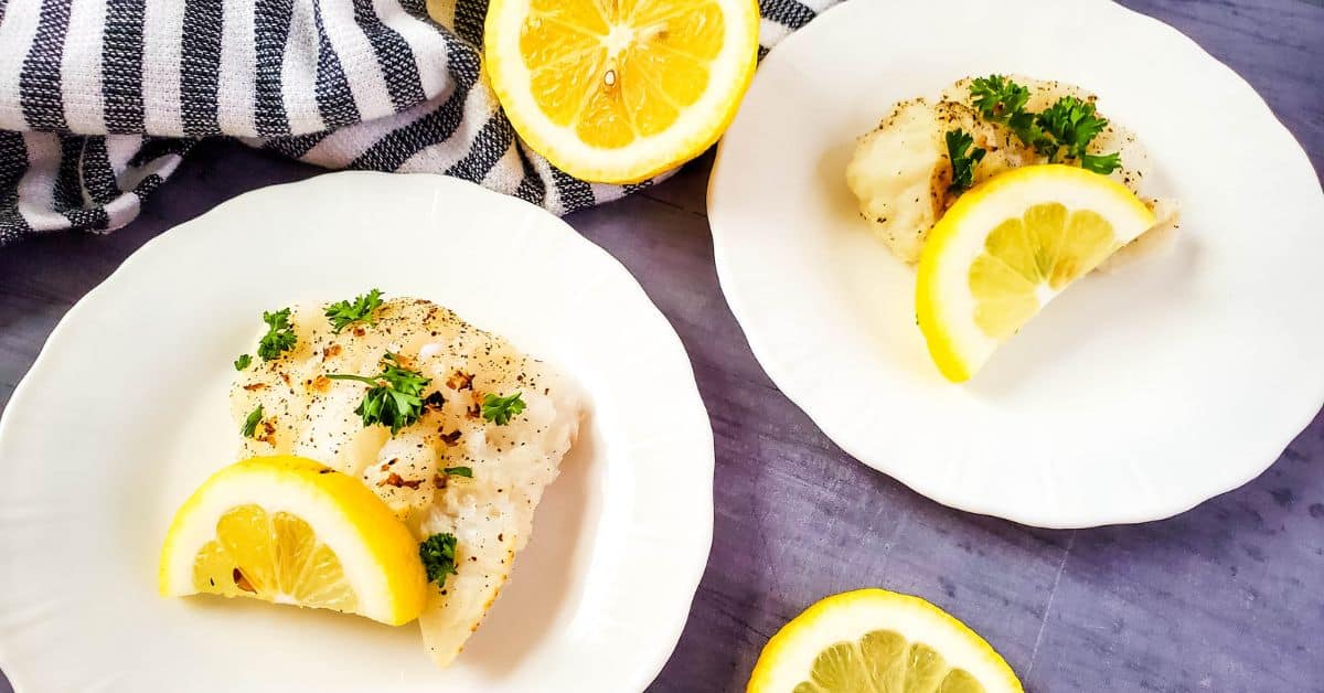 Two white plates with Air Fryer Cod fillets, garnished with lemon slices and parsley, on a gray surface with a striped cloth and additional lemon slices.