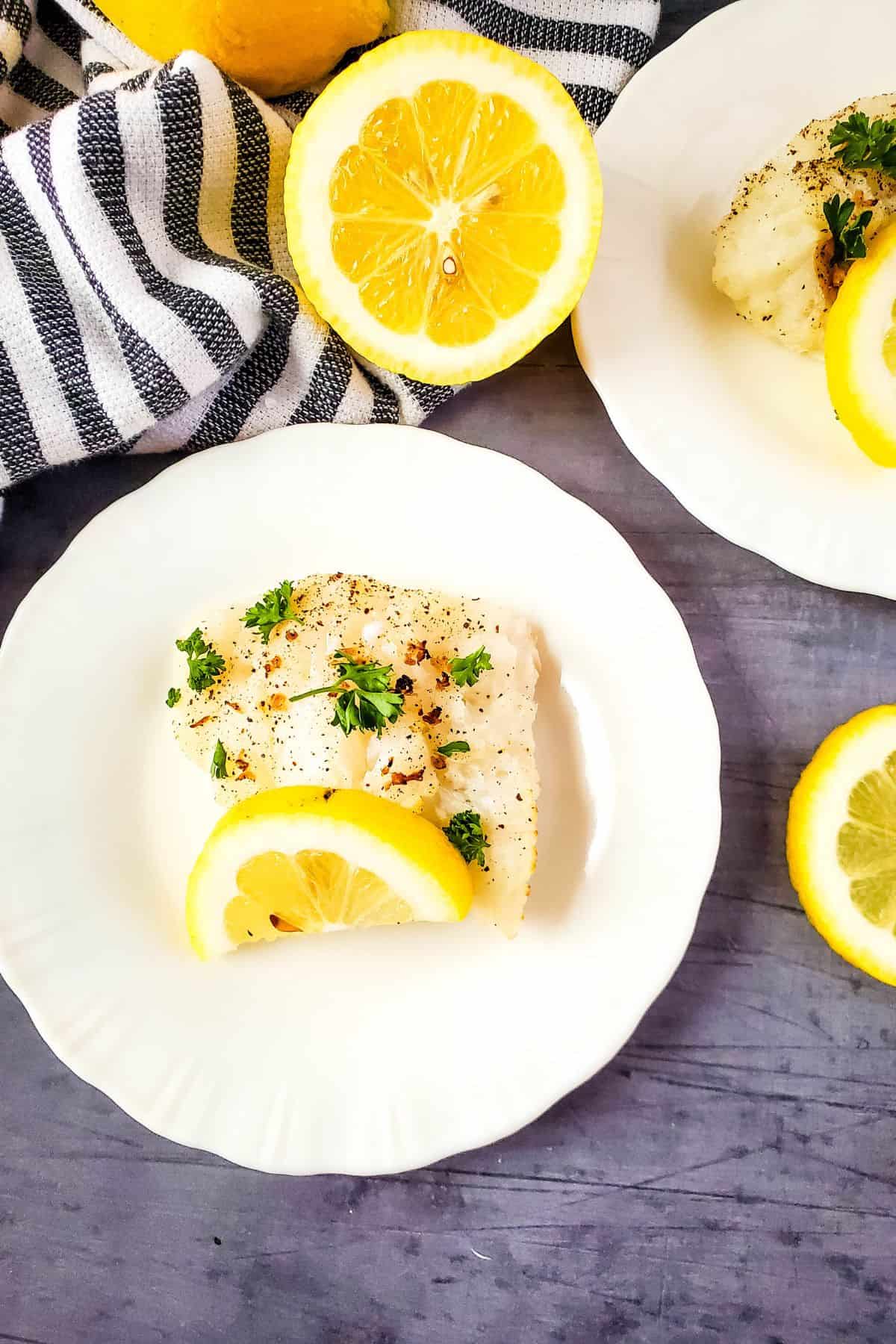 A piece of baked white fish, prepared as Air Fryer Cod and garnished with parsley and a lemon slice, is served on a white plate next to a halved lemon and a striped cloth.
