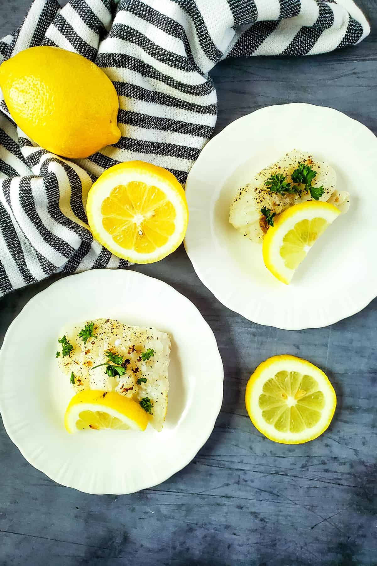Two white plates with seasoned Air Fryer Cod fillets garnished with parsley and lemon slices, next to a whole lemon and a striped cloth on a gray surface.