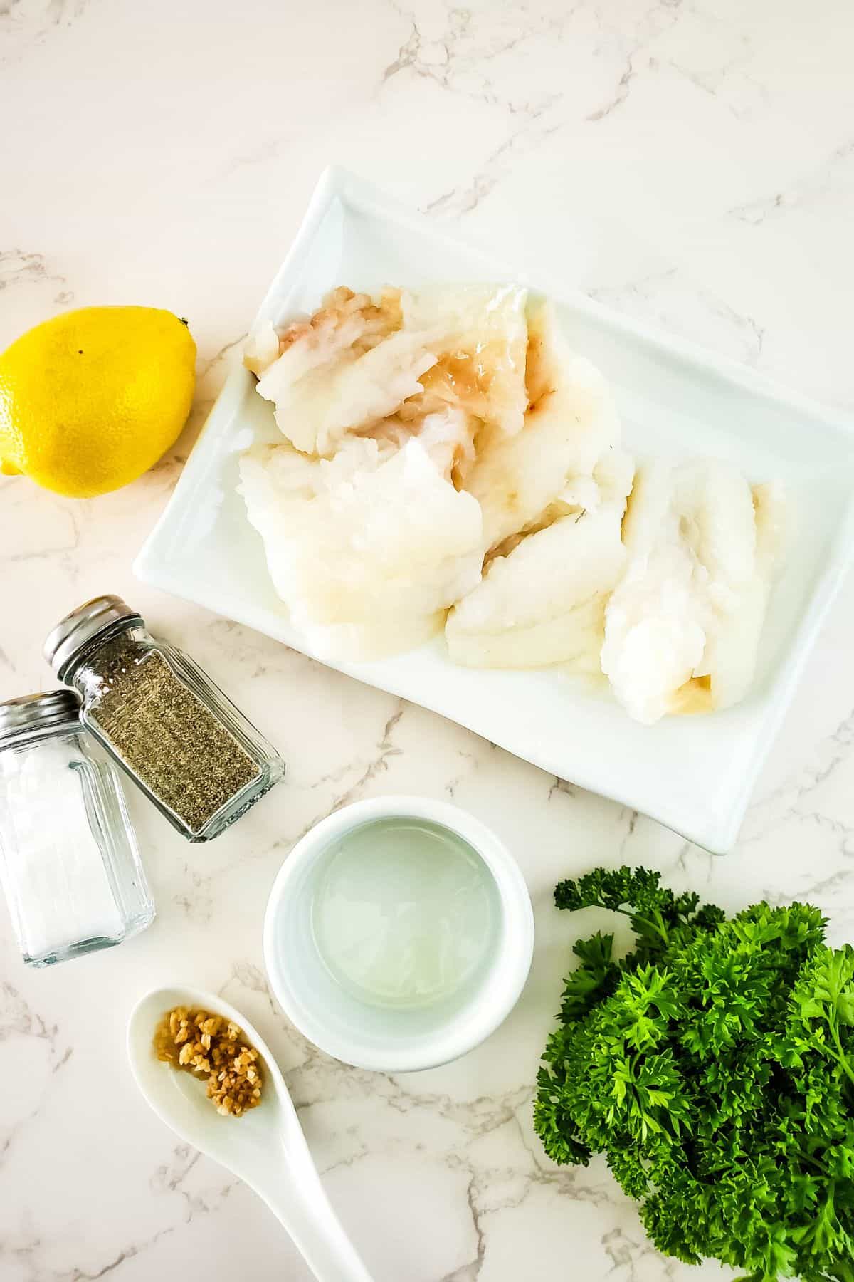 Raw white fish fillets on a white plate with a lemon, salt and pepper shakers, chopped parsley, minced garlic, and a small bowl of oil on a marble surface—everything you need to prepare delicious Air Fryer Cod.