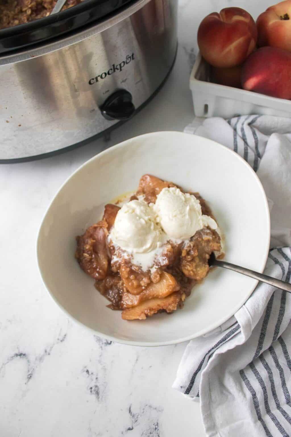 A bowl of crock pot peach cobbler topped with two scoops of vanilla ice cream sits next to a slow cooker and a box of fresh peaches on a white kitchen counter.