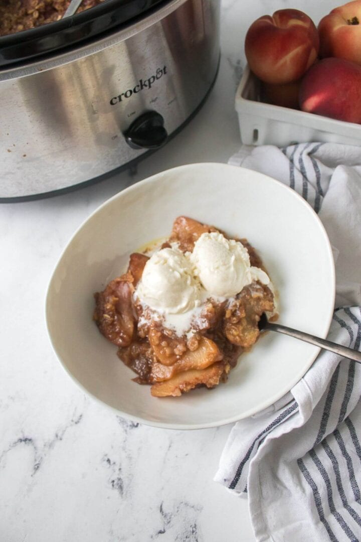 A bowl of crock pot peach cobbler topped with two scoops of vanilla ice cream sits next to a slow cooker and a box of fresh peaches on a white kitchen counter.