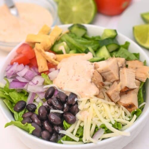A burrito bowl with lettuce, black beans, grilled chicken, shredded cheese, onions, tomatoes, tortilla strips, cucumbers, and creamy dressing—a delicious twist on a Spicy Southwest Salad—served with lime and tomato in the background.