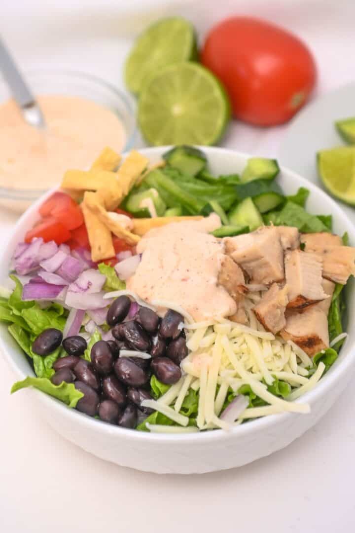 A burrito bowl with lettuce, black beans, grilled chicken, shredded cheese, onions, tomatoes, tortilla strips, cucumbers, and creamy dressing—a delicious twist on a Spicy Southwest Salad—served with lime and tomato in the background.