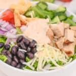 A bowl with chopped lettuce, grilled chicken, black beans, shredded cheese, onions, tomatoes, cucumbers, tortilla strips, and creamy dressing comes together to create a flavorful Spicy Southwest Salad, with lime wedges on a plate in the background.