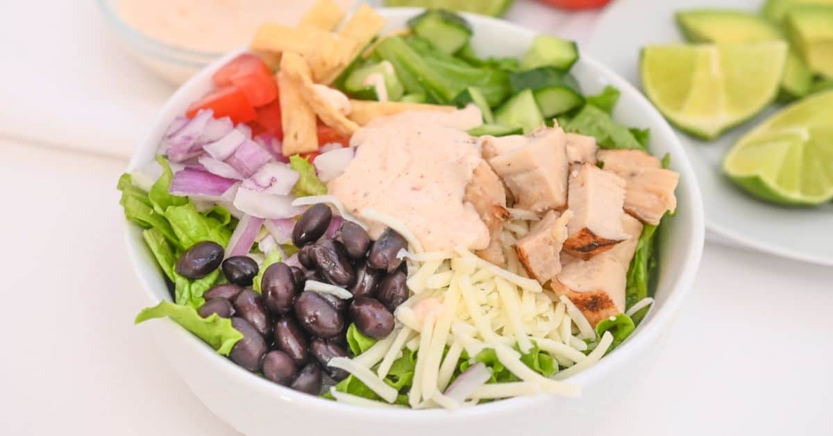 A bowl with chopped lettuce, grilled chicken, black beans, shredded cheese, onions, tomatoes, cucumbers, tortilla strips, and creamy dressing comes together to create a flavorful Spicy Southwest Salad, with lime wedges on a plate in the background.