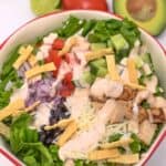 A bowl with lettuce, grilled chicken, black beans, shredded cheese, tortilla strips, diced tomato, red onion, cucumber, and creamy dressing, with avocado and lime in the background.