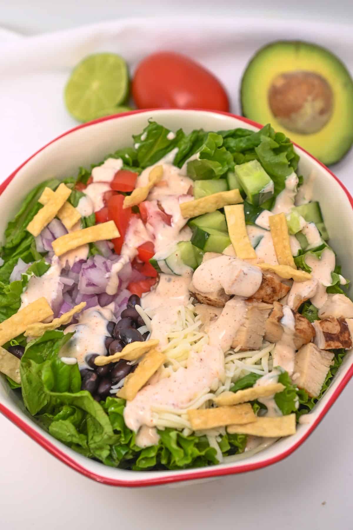 A bowl with lettuce, grilled chicken, black beans, shredded cheese, tortilla strips, diced tomato, red onion, cucumber, and creamy dressing, with avocado and lime in the background.
