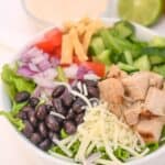 A bowl of Spicy Southwest Salad featuring crisp lettuce, black beans, shredded cheese, grilled chicken, diced tomatoes, red onions, cucumber, and crunchy tortilla strips sits front and center, with a bowl of dressing and lime halves in the background.